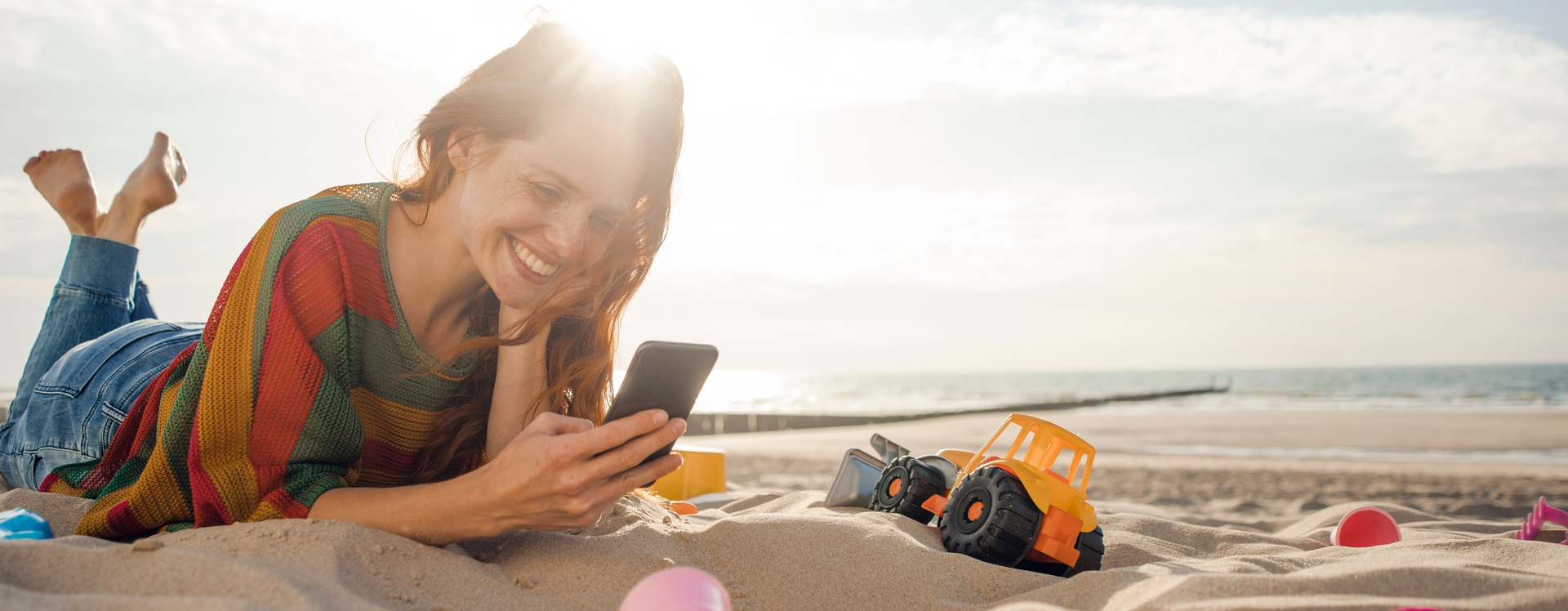 A woman is lying on the beach, smiling while looking at her smartphone IFB